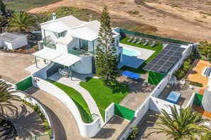 Vista aérea de un moderno edificio de dos plantas con una entrada de vehículos curva, una piscina y paneles solares.