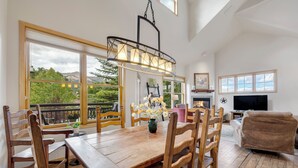 A dining area with a wooden table and chairs, a hanging light fixture, and large windows with a view of greenery and mountains. - Stunning 3-Bedroom Top Floor Condo – Walk to Lifts and Downtown (Breckenridge)