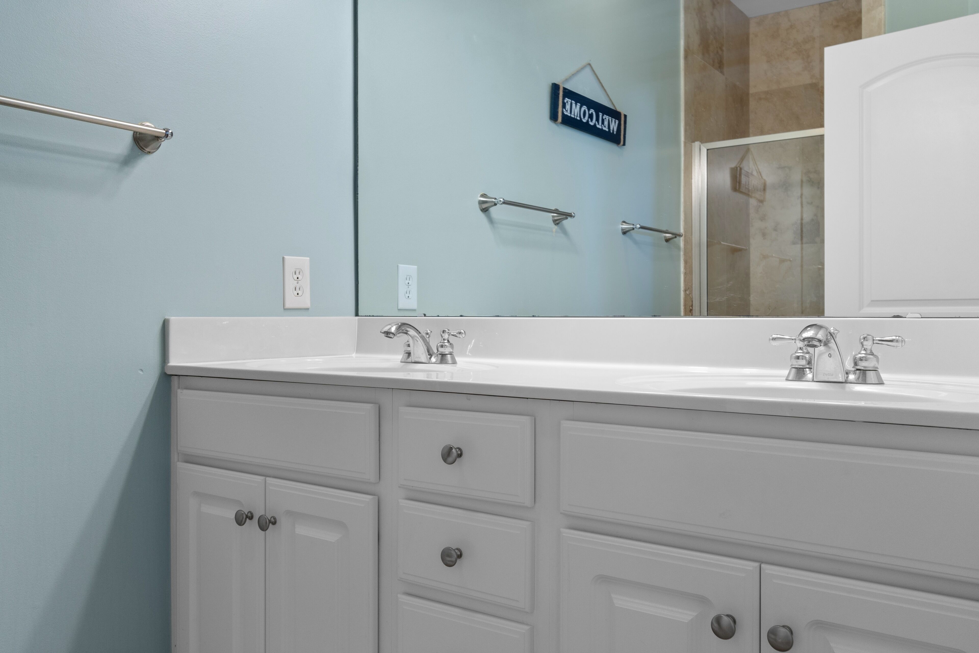 A bathroom with a white vanity, two sinks, a towel rack, and a welcome sign.