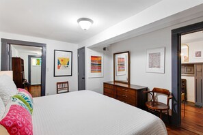Ein Schlafzimmer mit einem großen Bett, einem Schminktisch und einem Stuhl. An den Wänden befinden sich gerahmte Kunstwerke.