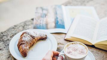 Una mano sosteniendo una cuchara sobre una taza de café, con un pastel en un plato, un libro abierto y un álbum de fotos sobre una mesa.
