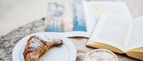 A hand holding a spoon over a cup of coffee with a pastry on a plate, with an open book and a photo album on a table.