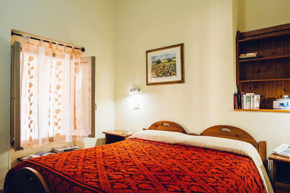 A bedroom with a bed, a nightstand, a wooden bookshelf, a window with sheer curtains, and a wall-mounted picture.