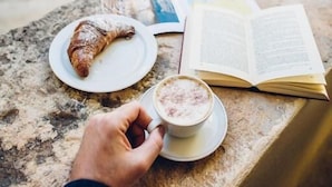 A person holding a cup of coffee, a croissant, and an open book on a stone table in an outdoor setting with arches in the background. - San Francesco Heritage Hotel (Alghero)
