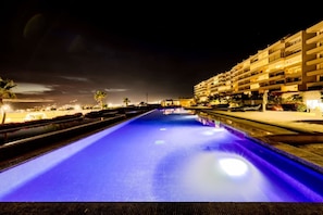 A nighttime view of a swimming pool with a blue-lit water surface, surrounded by a multi-story hotel building with balconies.