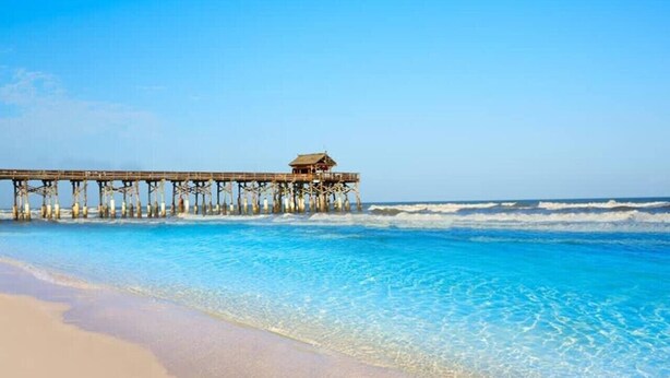 Une jetée s’avance dans une eau d’un bleu limpide, avec une plage de sable en premier plan.