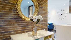Un espace d’accueil avec un mur en briques, un grand miroir décoratif, un vase rempli de fleurs blanches, une horloge et un fauteuil bleu.