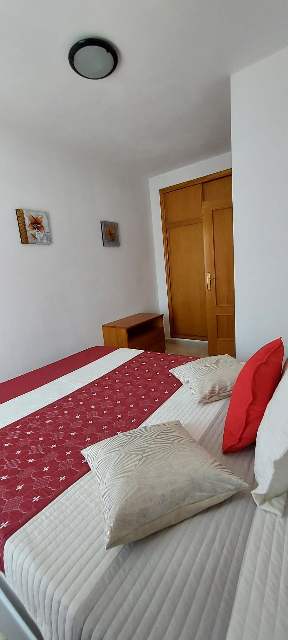 A bedroom with a bed, a wooden wardrobe, a small table, and two wall-mounted pictures.