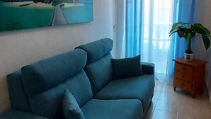 A living room with a blue sofa, a painting of a beach scene, a potted plant, and a window with blue curtains.