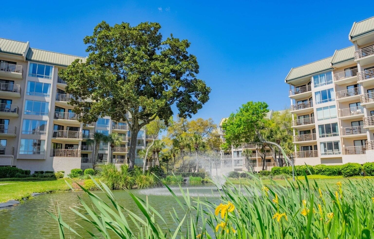 Un moderno edificio de apartamentos con varios balcones, una fuente central y abundante vegetación.