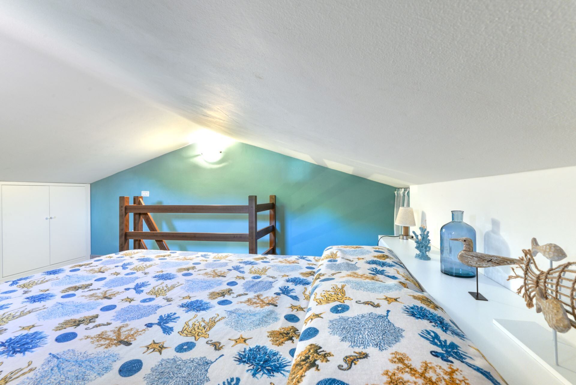 A bedroom with a sloped ceiling, a bunk bed, a wooden ladder, and a decorative wall with a blue and white pattern.