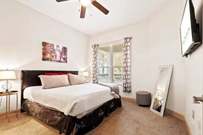Une chambre à coucher comprenant un lit, deux tables de chevet, un ventilateur de plafond, une fenêtre avec des rideaux, un téléviseur fixé au mur et un tableau encadré.