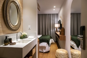 Una camera d'albergo moderna con un grande specchio, un lavandino bianco, un comodino con una lampada e due letti con coperte verdi.