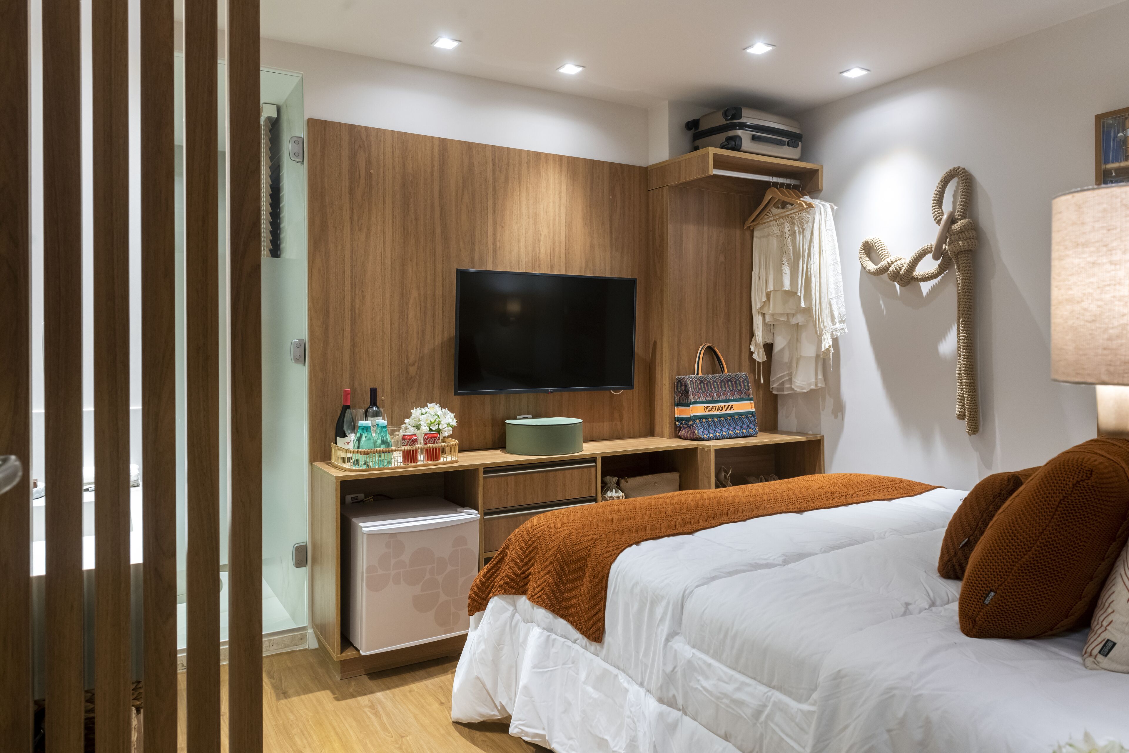 A modern bedroom with a bed, a TV, a small fridge, a wooden wardrobe, and a hanging coat rack.