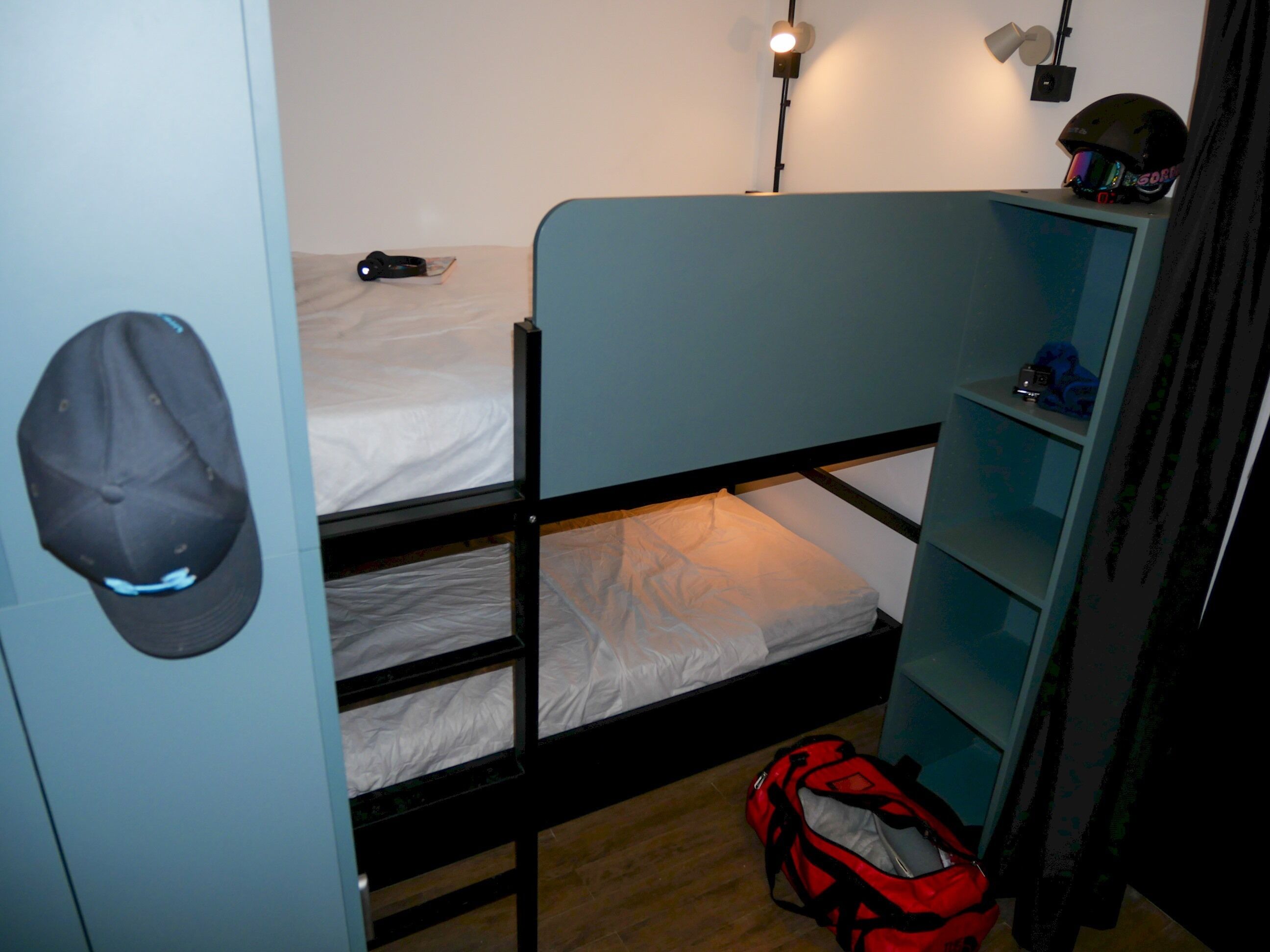 A bunk bed room with a single bed, a desk, a chair, a red backpack, and a helmet on the shelf.