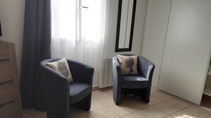 Two dark armchairs with patterned cushions in a room with tiled flooring, a window with sheer curtains, and a white wardrobe.