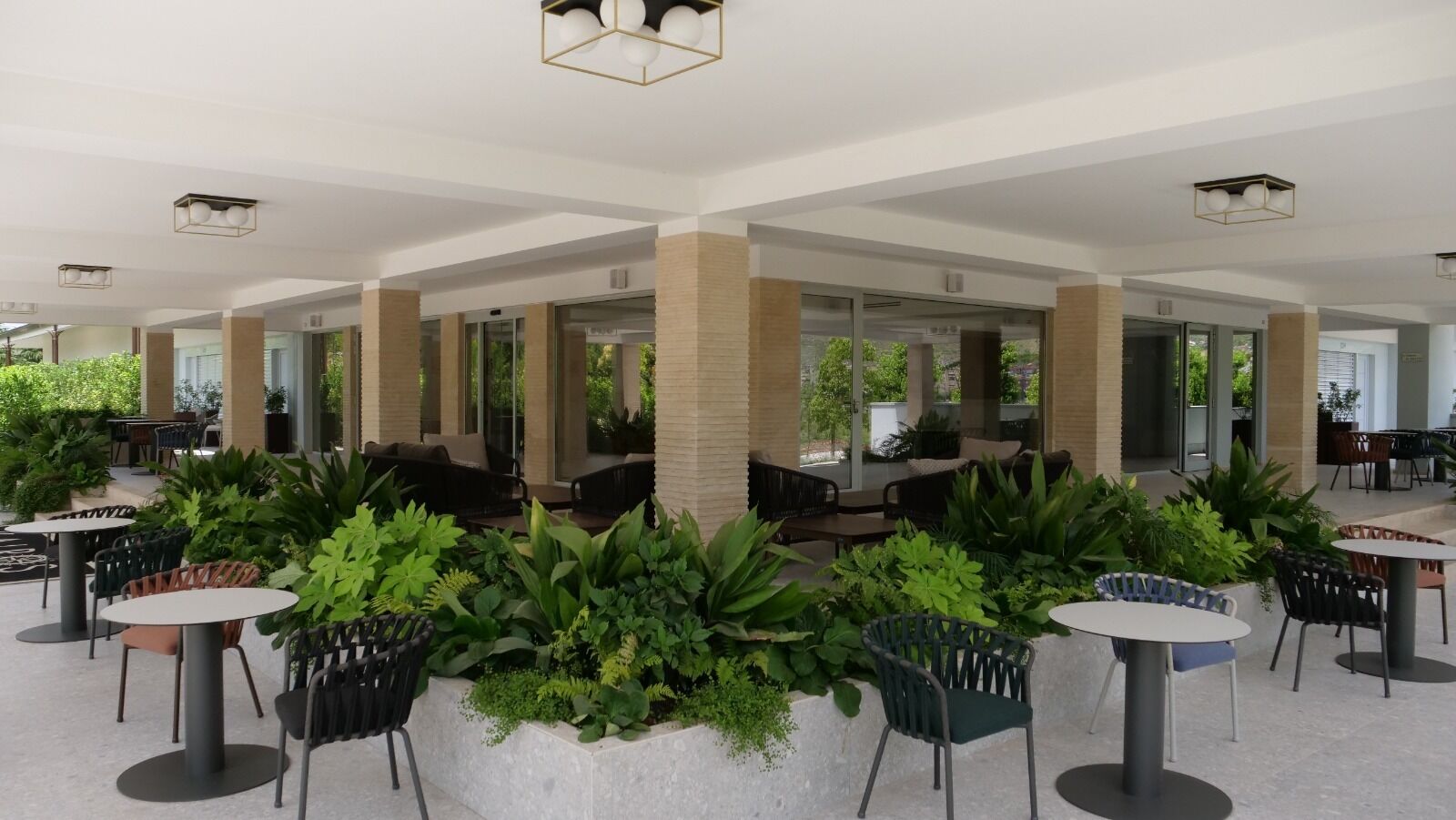 A spacious indoor area with multiple seating arrangements, including tables and chairs, surrounded by lush green plants.