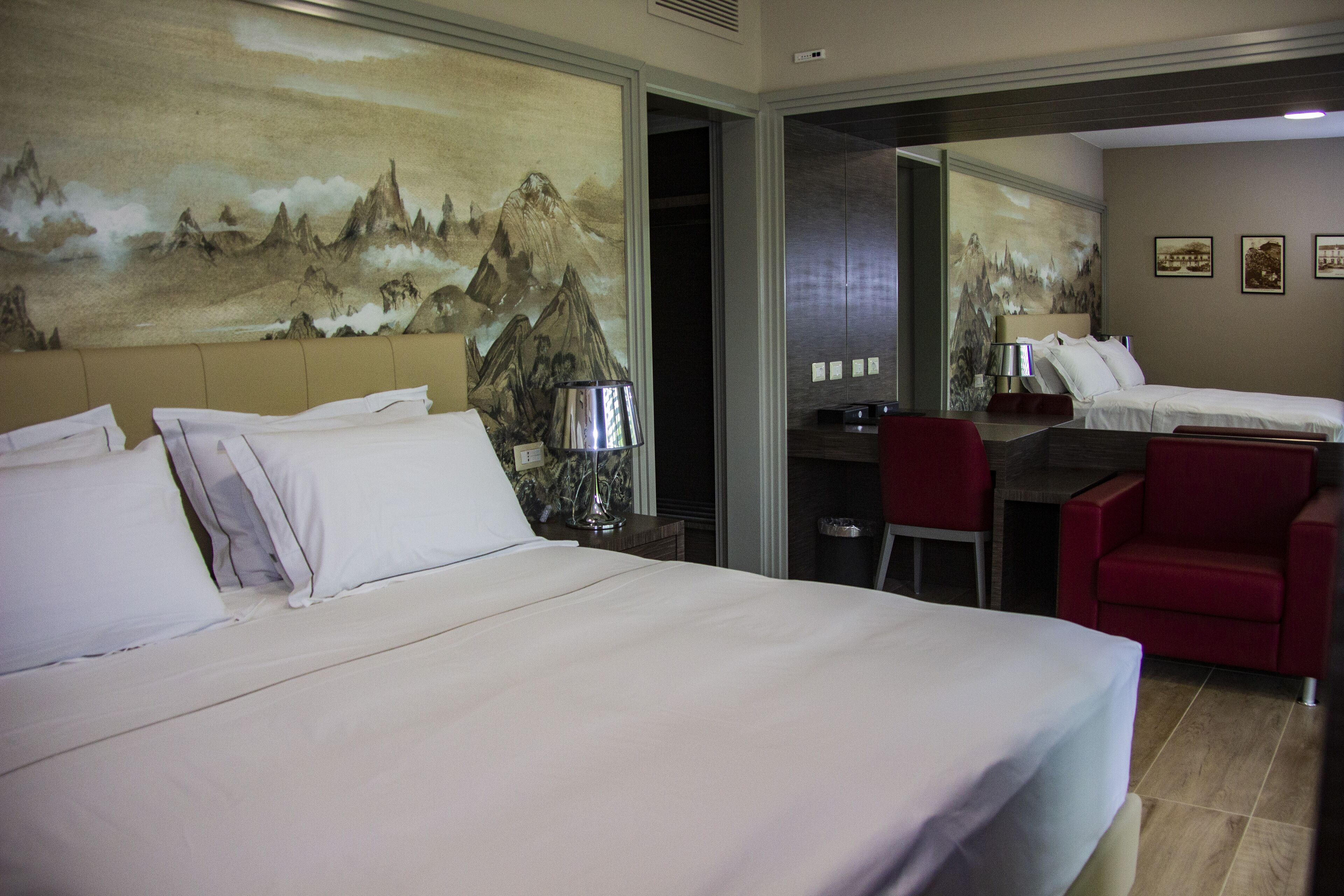 Une chambre d’hôtel avec un grand lit, un bureau, une chaise et un fauteuil rouge. Il y a un tableau au mur et un miroir qui reflète la pièce.