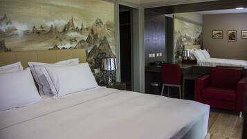A hotel room with a large bed, a desk, a chair, and a red armchair. There is a painting on the wall and a mirror reflecting the room.