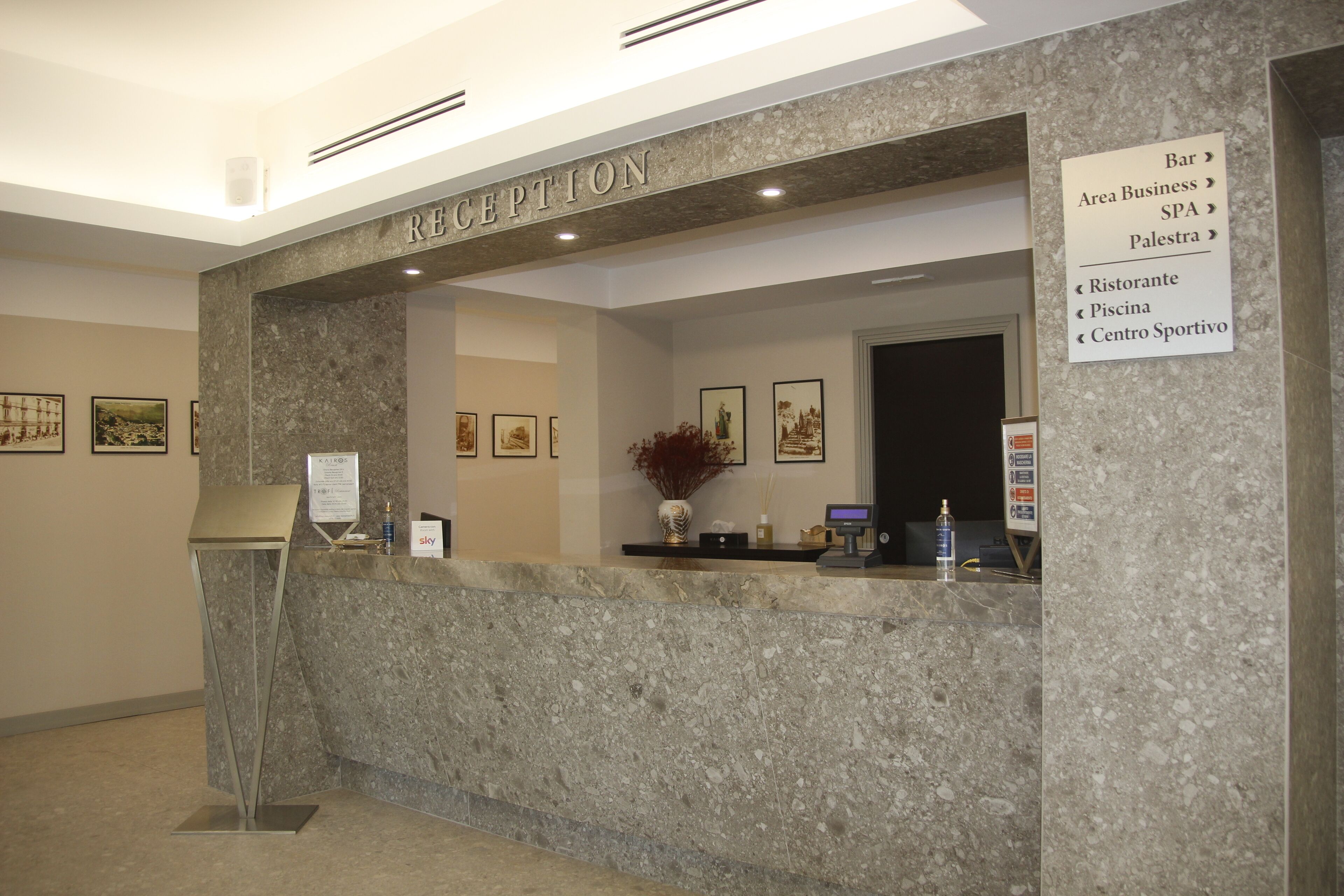 A reception area with a marble counter, a reception desk, framed artwork on the walls, and a sign listing hotel amenities.
