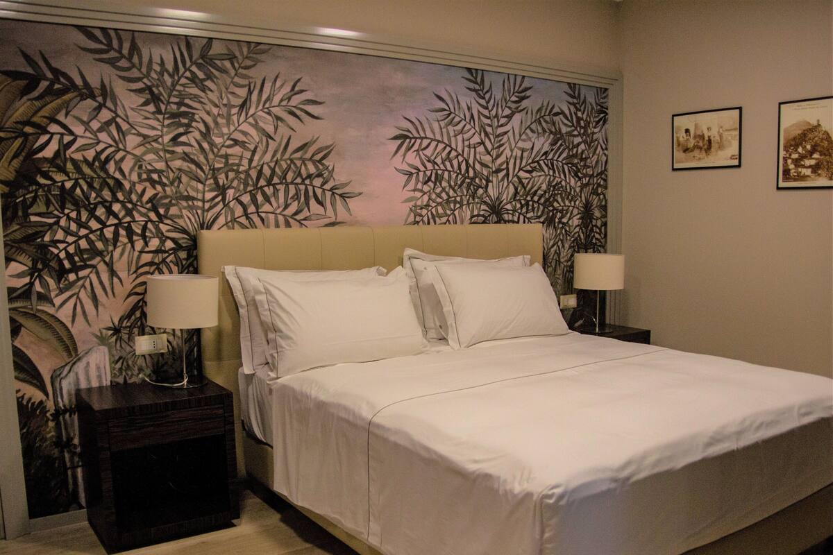 A neatly made bed with white linens, two bedside lamps, and a wall with a large botanical print.
