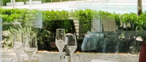 Mesa posta para refeição, com copos transparentes e um vaso, com vista para um jardim bem cuidado e uma piscina.