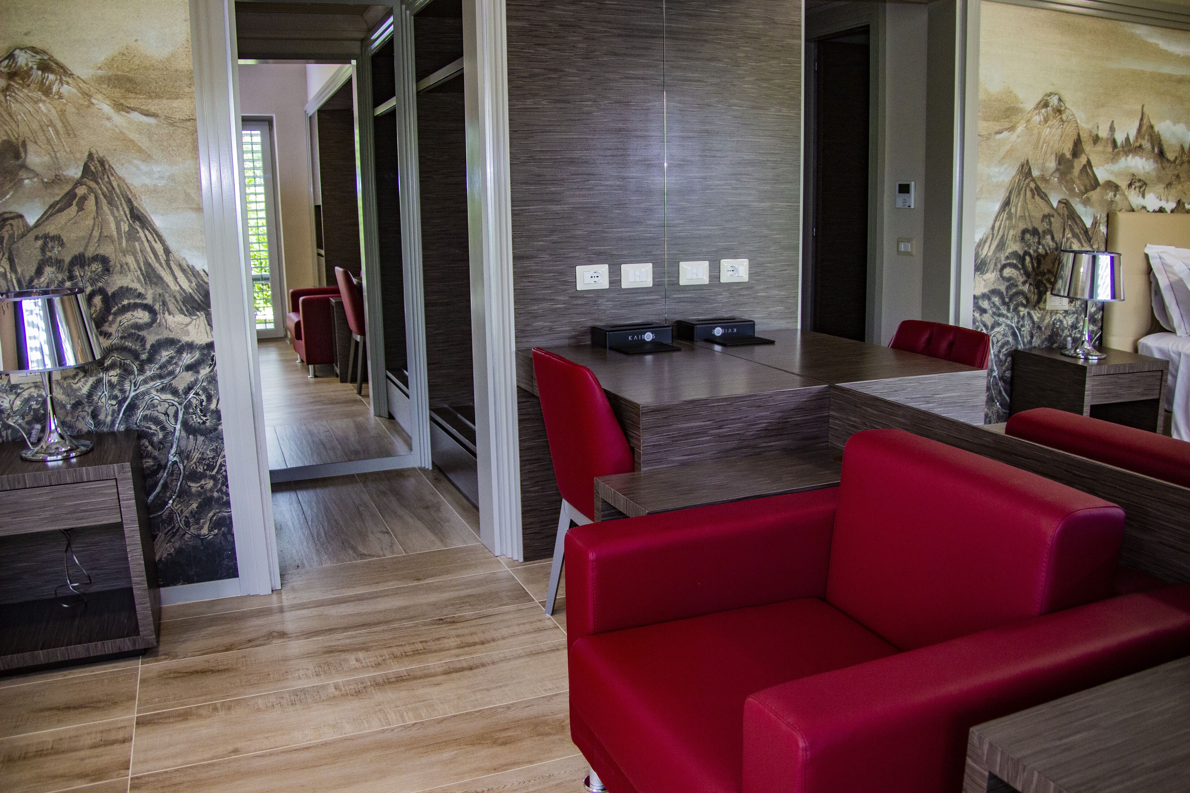 A modern hotel room with a wooden floor, red leather sofa, a dining table with chairs, and a painting of mountains on the wall.