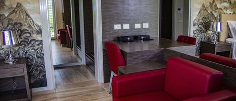Una habitación de hotel moderna con piso de madera, sofá de cuero rojo, una mesa de comedor con sillas y un cuadro de montañas en la pared.