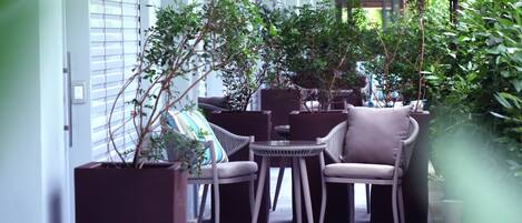A cozy outdoor seating area with wicker furniture, potted plants, and a view of the building's interior.