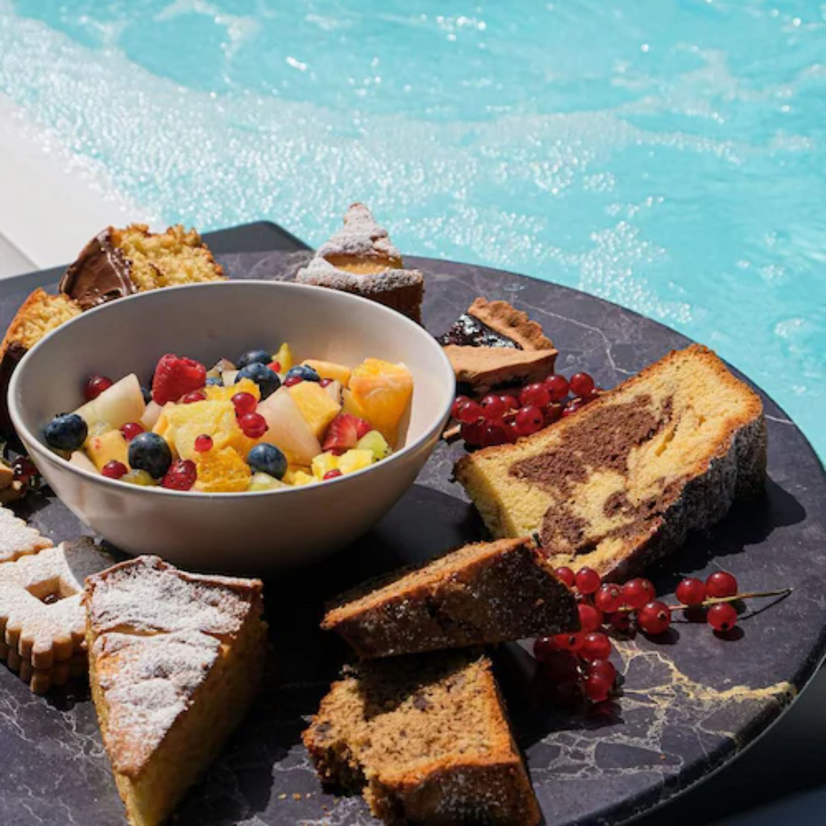 A breakfast spread with a bowl of fruit salad, various pastries, and a slice of cake on a dark plate by a pool.