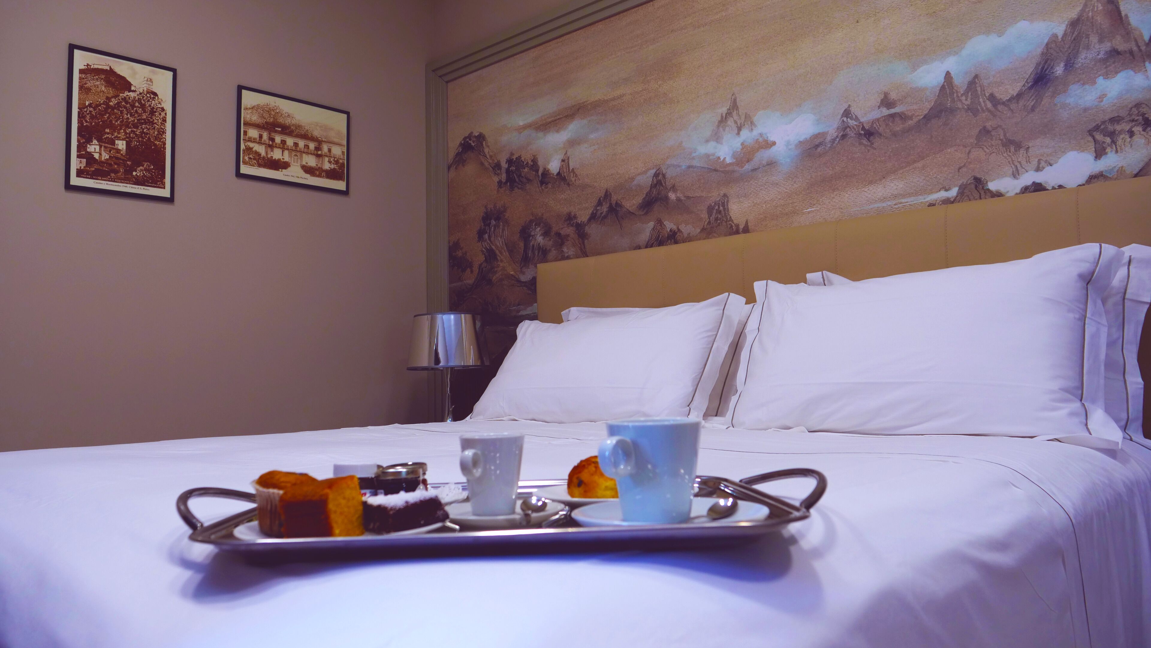 Une chambre d’hôtel avec un lit, un plateau avec de la nourriture et des boissons, et deux tableaux encadrés accrochés au mur.