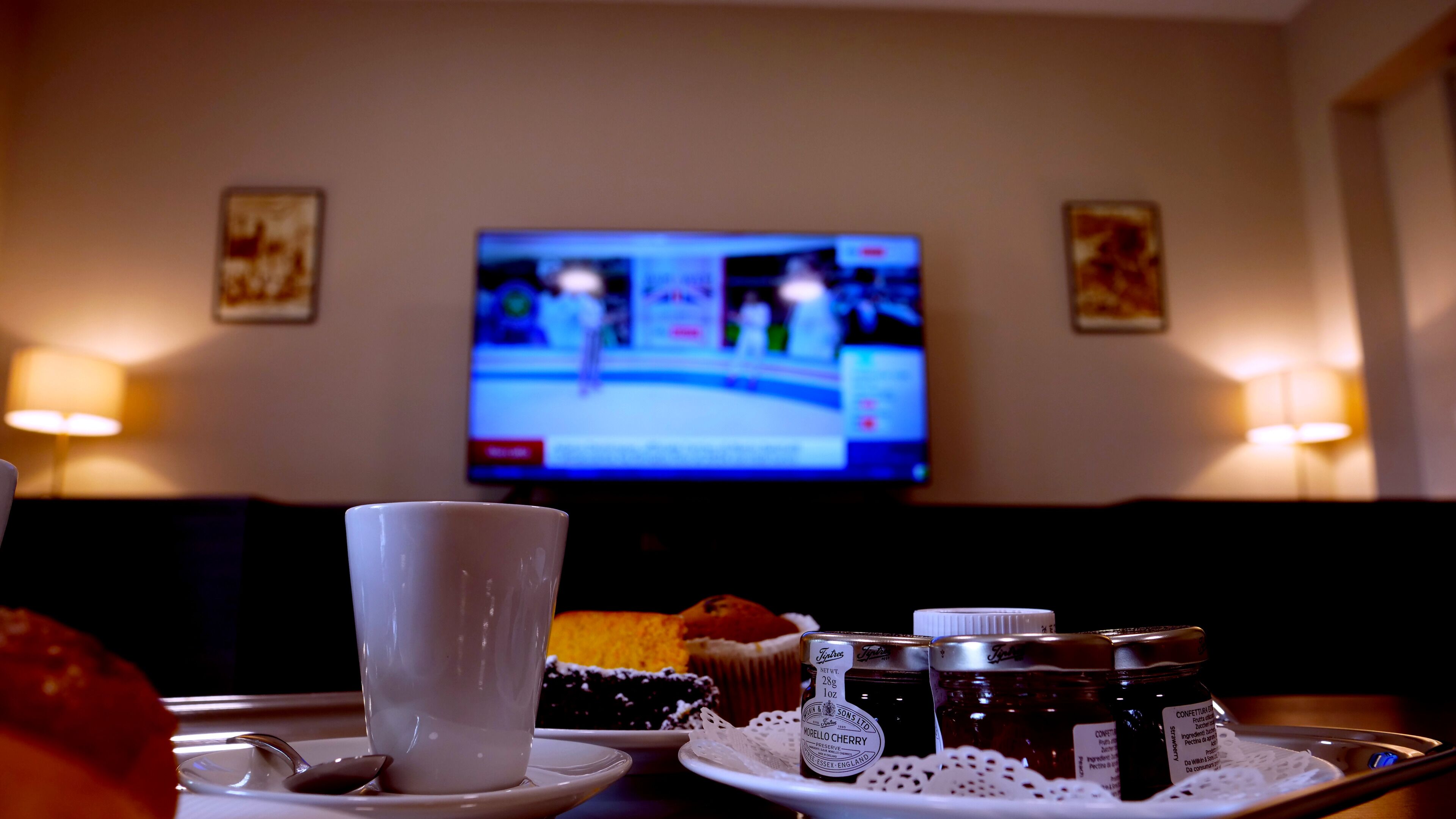 Zona pranzo con una televisione che mostra un evento sportivo, una tazza bianca su un piattino, un vassoio con cibo e condimenti e un portacandele.