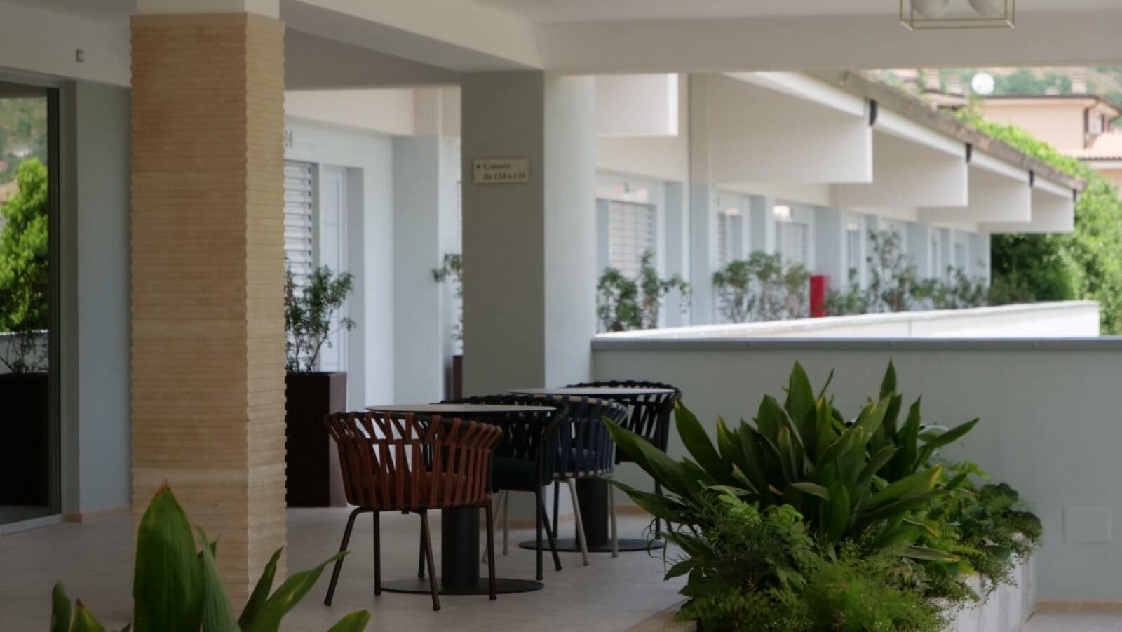 A covered outdoor seating area with tables and chairs, surrounded by potted plants and a white wall.