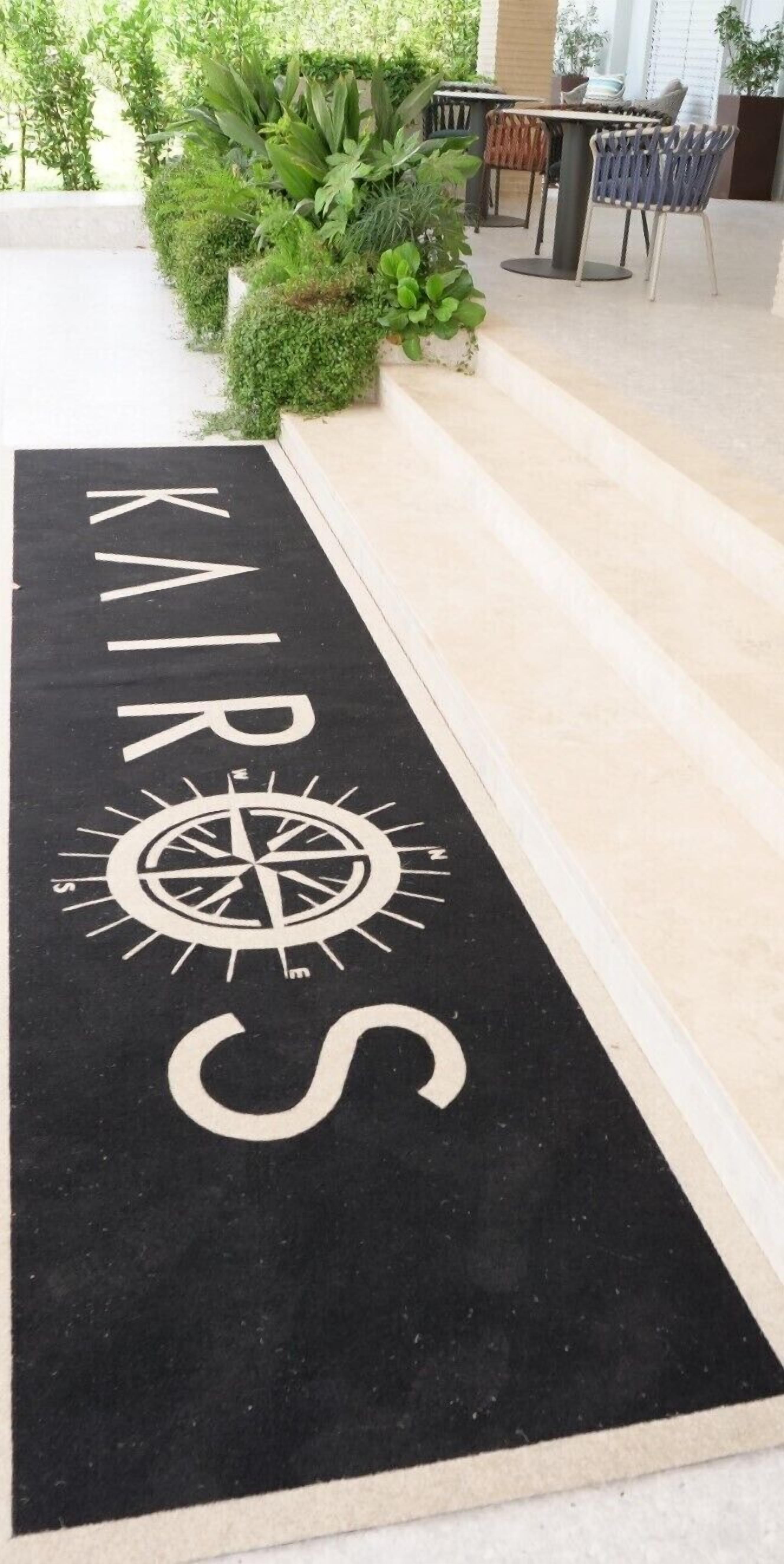 Un felpudo negro con la palabra "KAROS" y un diseño de brújula, que conduce a una escalera con mobiliario exterior.