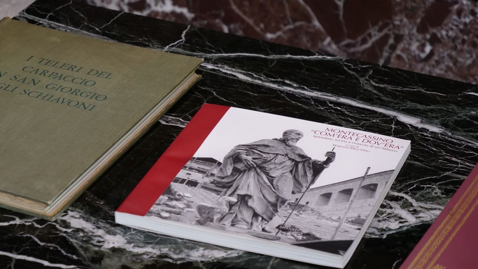 Dos libros, uno titulado "I Teleri del Carpaccio" y otro "Monte Cassino 'Comera Dovèra'", sobre una superficie de mármol.