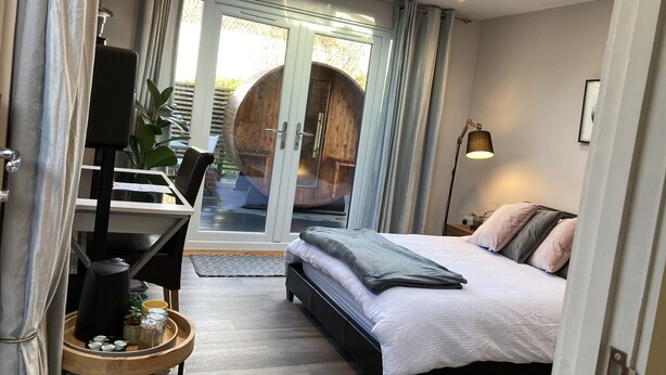 A bedroom with a bed, a desk with a chair, a lamp, and a large wooden barrel outside the window. - Contemporary Guest House With Outside Barrel Sauna (Wadebridge)