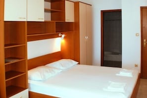 Une chambre avec une armoire en bois, un lit avec une literie blanche et une porte surmontée d’un rideau foncé.