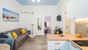Ein modernes Wohnzimmer mit einem grauen Sofa, einem kleinen runden Tisch und einem Holzesstisch. Ein Küchenbereich mit Arbeitsplatte aus Holz und einer Spüle ist zu sehen.