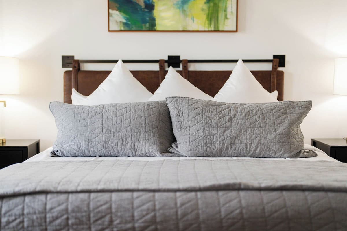Una cama con colcha gris y dos almohadas grises, cabecera de madera y un cuadro en la pared.
