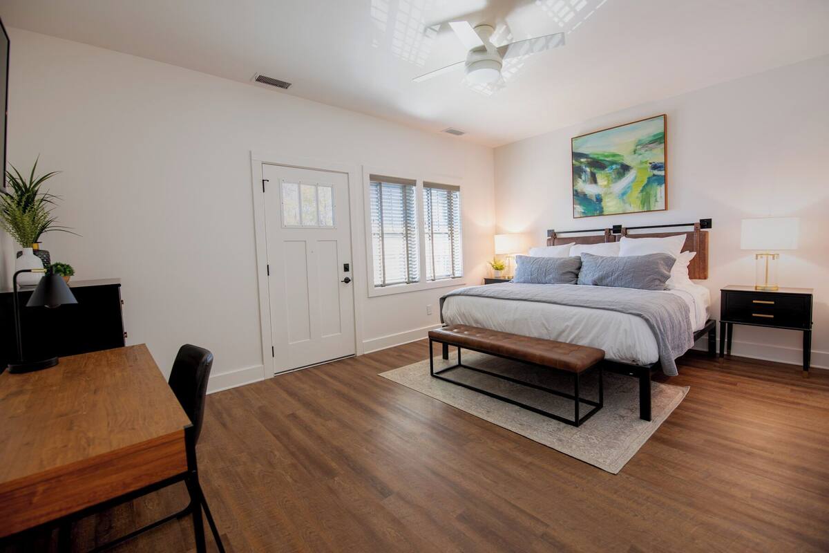 Un dormitorio con cama, mesitas de noche, escritorio, silla, banco y una puerta.