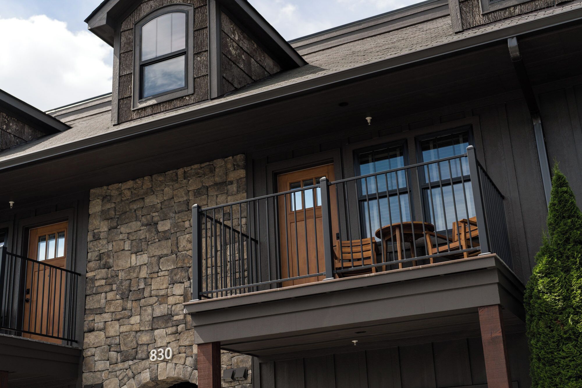 A two-story building with a stone facade, a balcony with wooden furniture, and a large window.