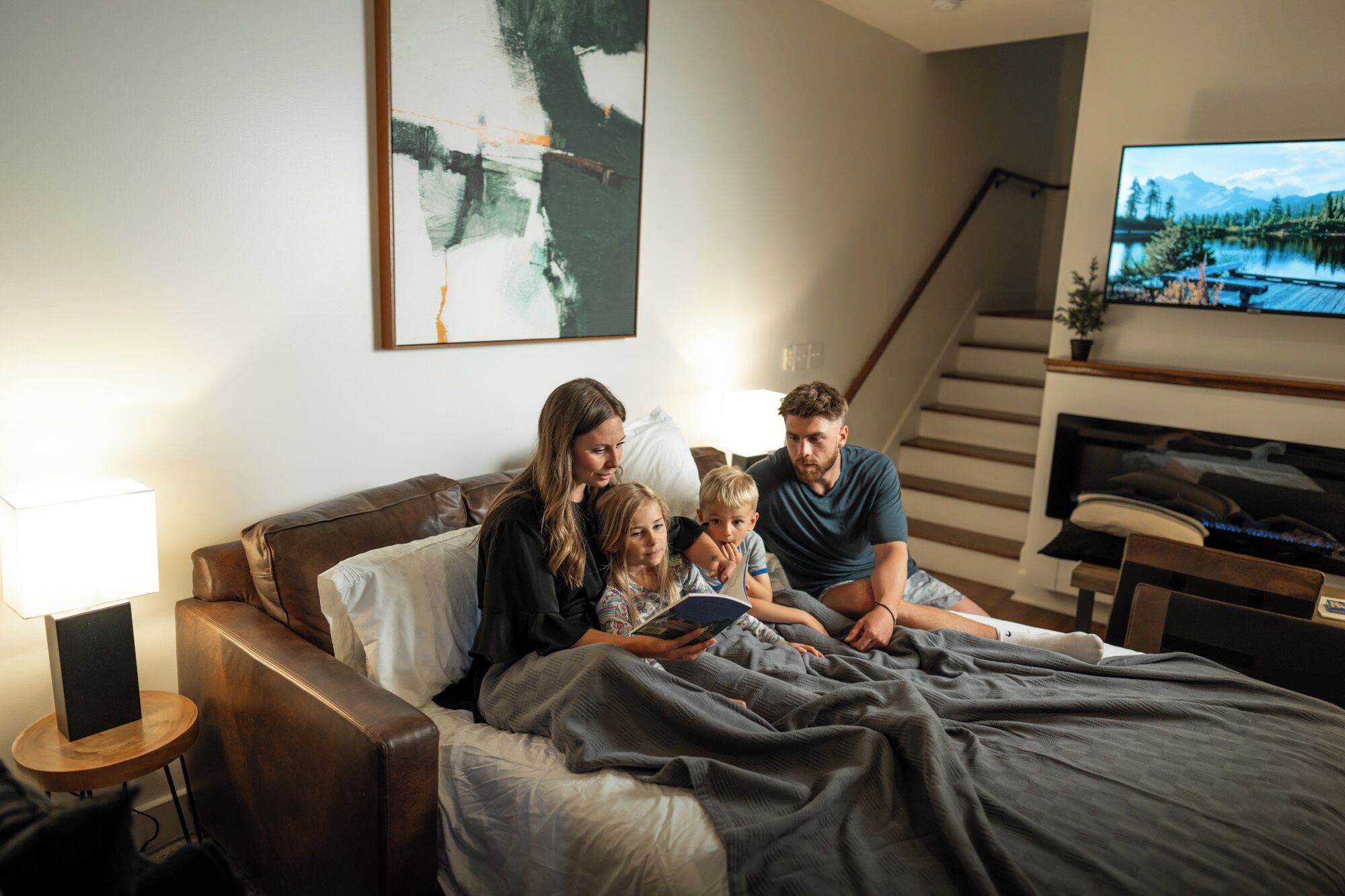 Una familia de cuatro personas en una cama, con un cuadro encima, un televisor en la pared y una escalera al fondo.