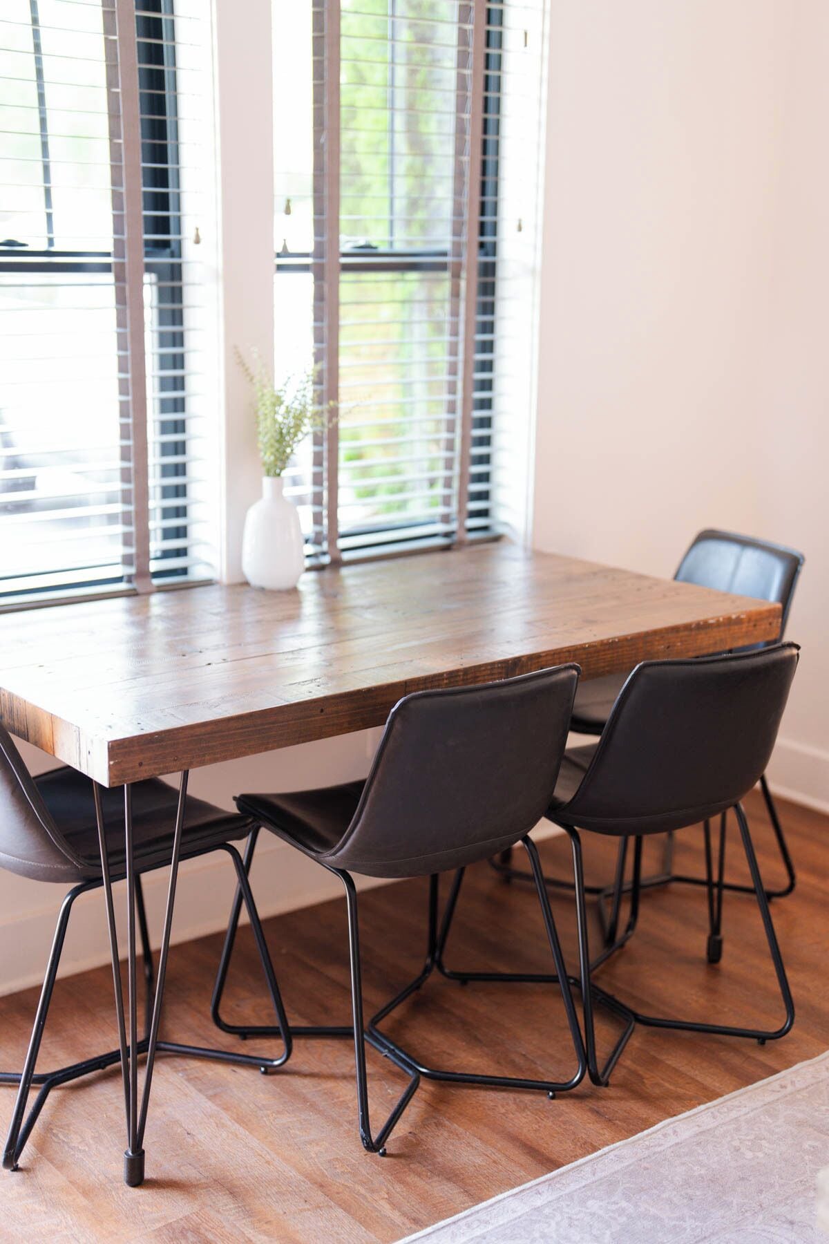 A dining table with four chairs, a vase with flowers, and a window with blinds.