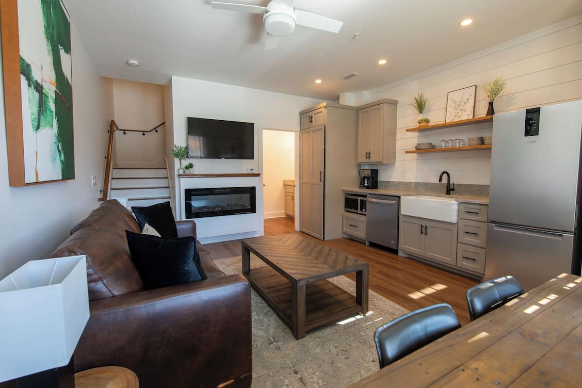 Una cocina moderna con horno empotrado, microondas y refrigerador. Hay un comedor con mesa y sillas de madera, y una sala de estar con sofá de cuero y una mesa de centro.