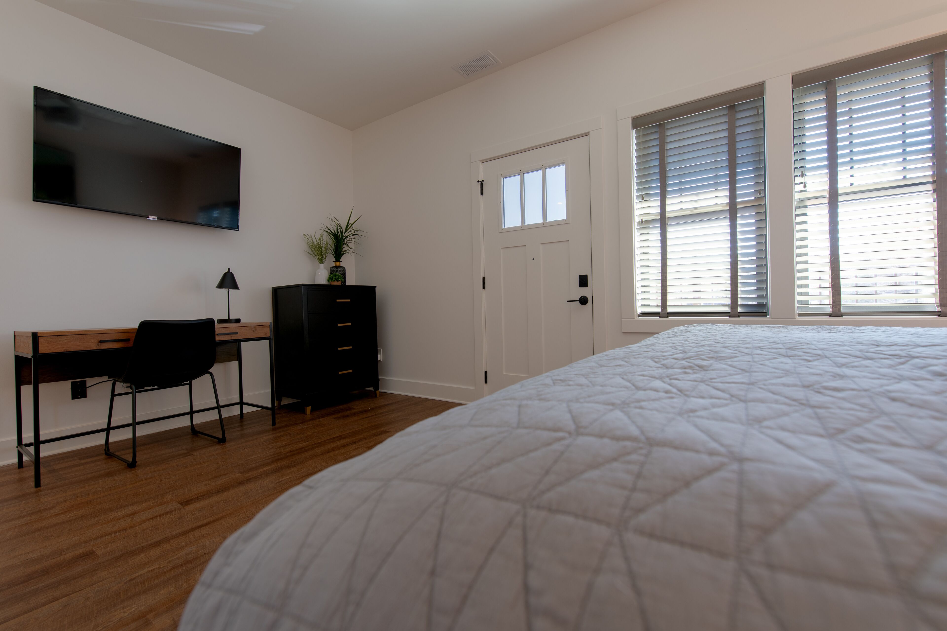 Un dormitorio con cama, escritorio, silla, televisor, cómoda y una puerta con ventanas.