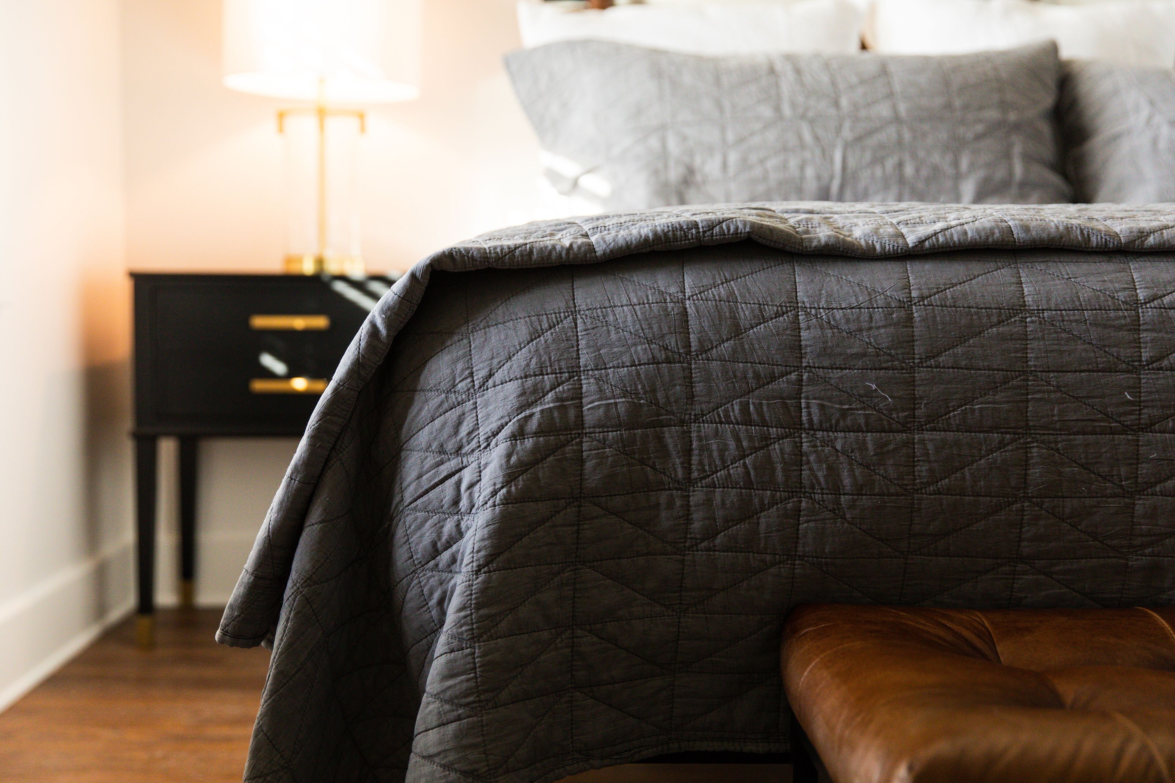 A neatly made bed with a grey quilt, a brown leather bench, and a nightstand with a lamp.