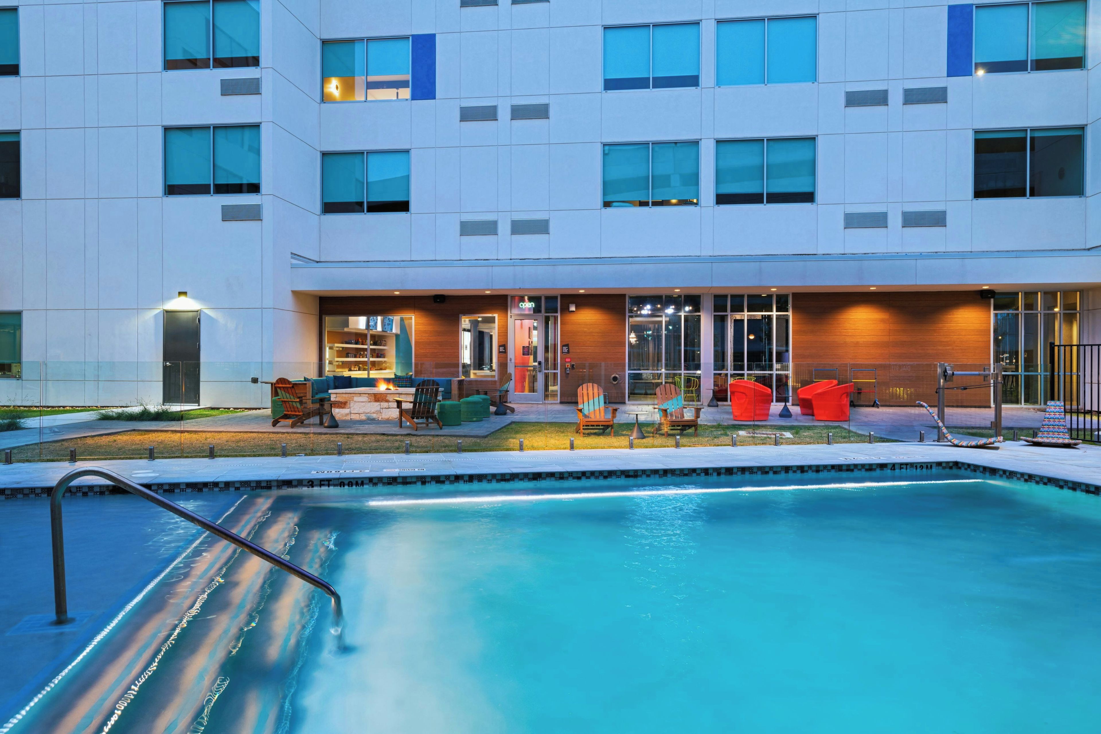 A modern building with a swimming pool, outdoor seating, and a well-lit entrance.
