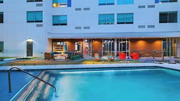 A modern building with a swimming pool, outdoor seating, and a well-lit entrance.