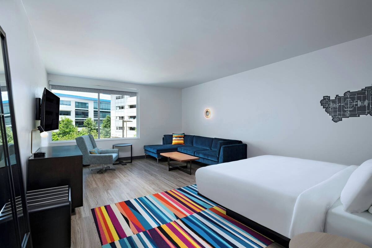 A modern hotel room with a large bed, a blue sofa, a small table, and a colorful striped rug.