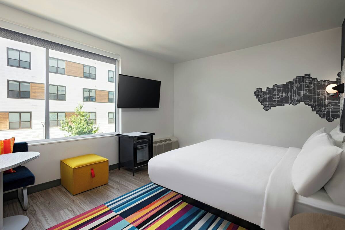 A hotel room with a bed, a desk, a chair, a small table, a colorful striped rug, a flat-screen TV, and a window with a view of a building.